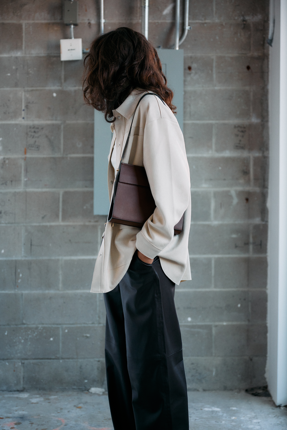 Model wearing the Low Rise bag in Cacao leather over the shoulder beside a brick wall