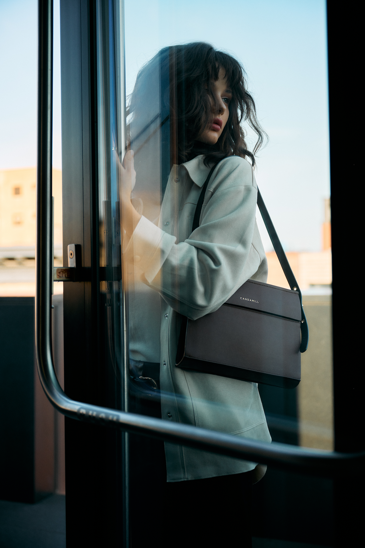 Model wearing the Low Rise bag in Cacao leather over the shoulder beside a glass door